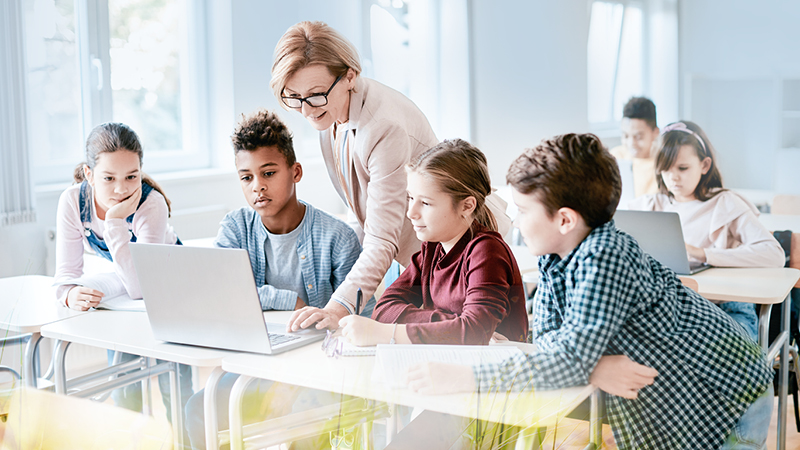 Eine Lehrerin sitzt mit einer Gruppe von Schulkindern in einem Klassenraum und zeigt ihnen auf einem Laptop etwas auf dem Bildschirm. Die Kinder schauen aufmerksam zu und versammeln sich gemeinsam um den Tisch.
