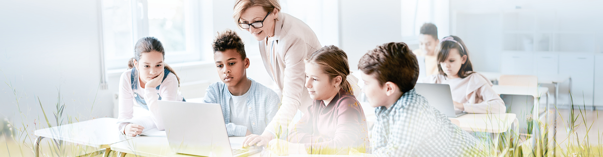 Headerbild, auf dem eine Lehrerin mit einer Gruppe von Schulkindern in einem Klassenraum steht und ihnen auf einem Laptop etwas auf dem Bildschirm zeigt. Die Kinder schauen aufmerksam zu und sind gemeinsam um den Tisch versammelt.